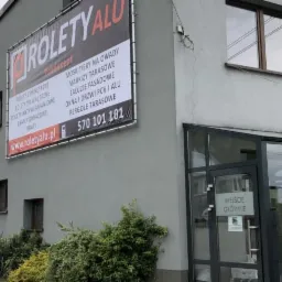 Budynek firmy z reklamą zewnętrzną oferującą rolety, moskitiery, markizy, żaluzje, okna, drzwi i bramy garażowe. Wejście główne do budynku przeszklone.