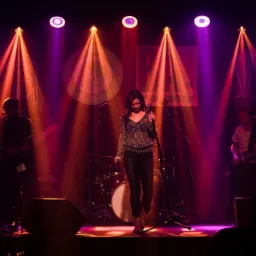 Wokalistka na scenie podczas koncertu, oświetlona reflektorami w odcieniach fioletu i pomarańczy, z gitarzystami po bokach i perkusją w tle.