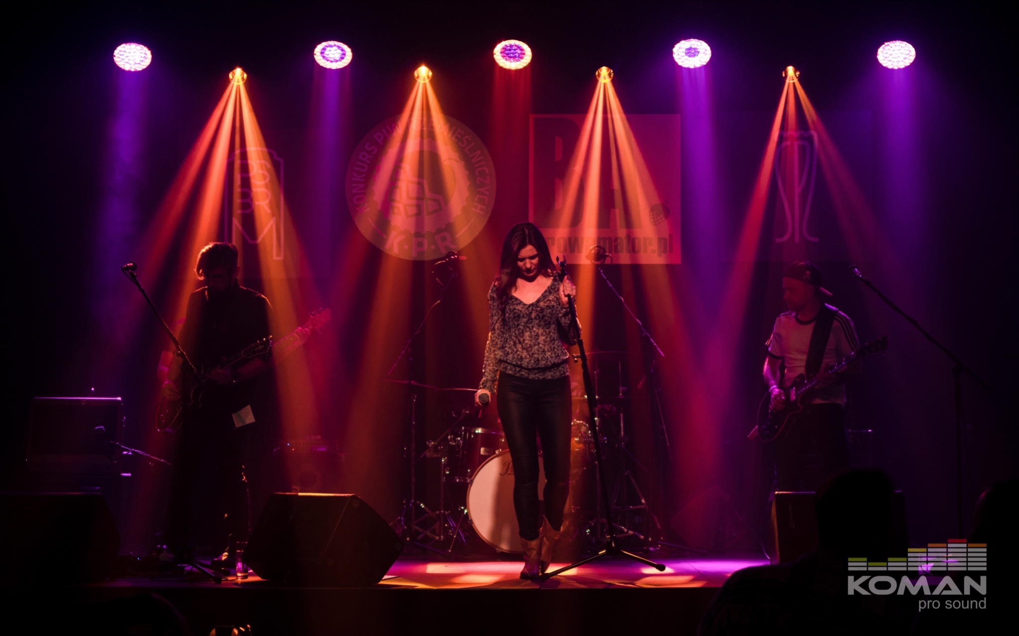 Wokalistka na scenie podczas koncertu, oświetlona reflektorami w odcieniach fioletu i pomarańczy, z gitarzystami po bokach i perkusją w tle.