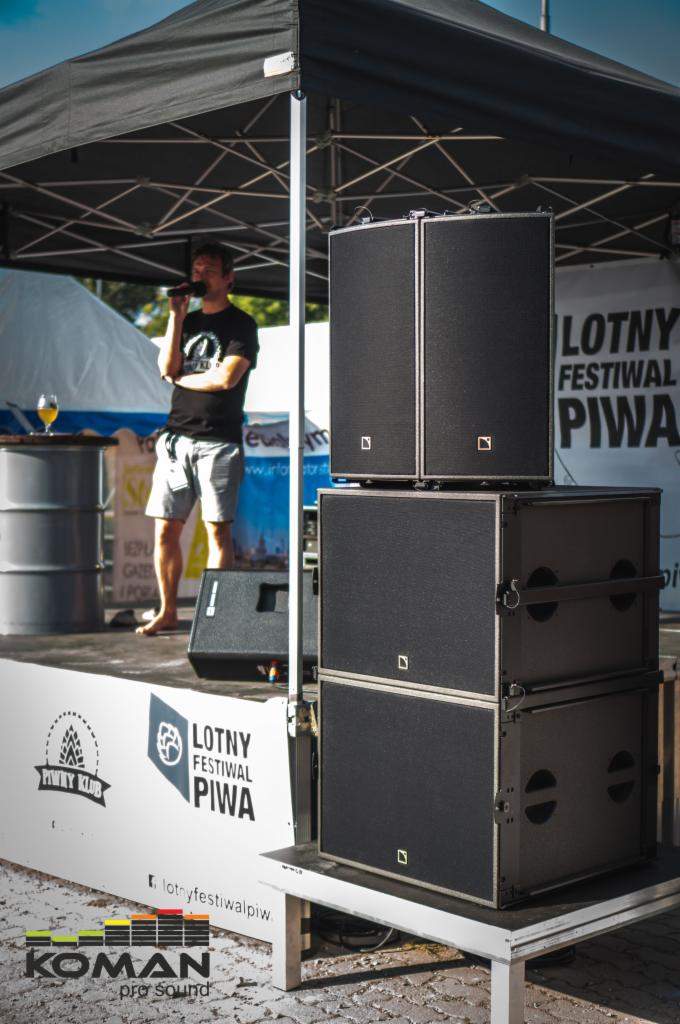 Scena plenerowa z ustawionymi kolumnami głośnikowymi i mężczyzną z mikrofonem podczas Lotnego Festiwalu Piwa.