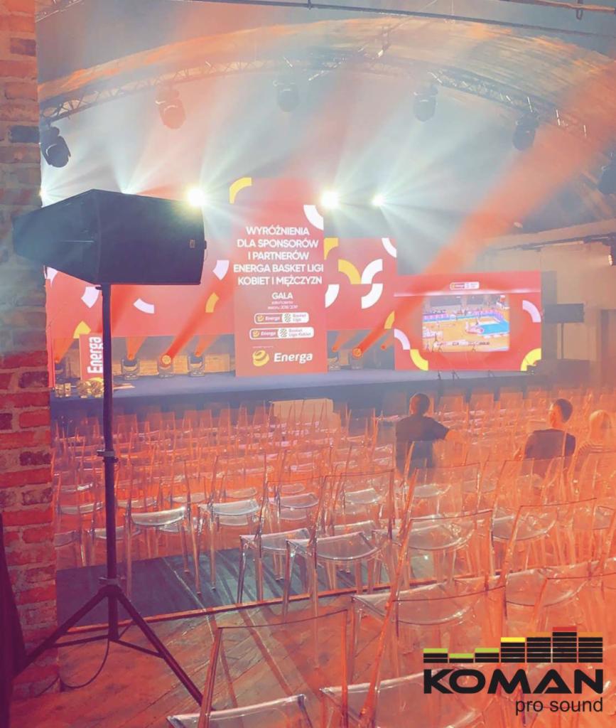 Sala konferencyjna z rzędami przezroczystych krzeseł, scena z telebimem wyświetlającym logo Energa Basket Ligi, profesjonalny głośnik na statywie z lewej strony, ceglana ściana w tle, oświetlenie...