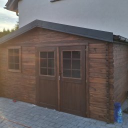 Aneta Deperas deperasdesign - Drewniana konstrukcja z dwuskrzydłowymi drzwiami i małym oknem, przylegająca do budynku z szarą elewacją, otoczona kostką brukową, z widocznymi elementami wyposażenia i butelką wody.