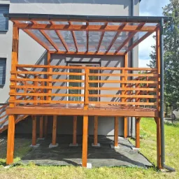 Drewniany taras z zadaszeniem z przezroczystego tworzywa, schody z balustradą, szare ściany budynku i zieleń drzew w tle, w słoneczny dzień.