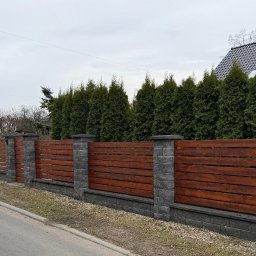 Aneta Deperas deperasdesign - Nowoczesne ogrodzenie z drewnianymi panelami i kamiennymi słupkami, otoczone zielenią, z domem w tle. Widok z poziomu ulicy, pochmurny dzień.