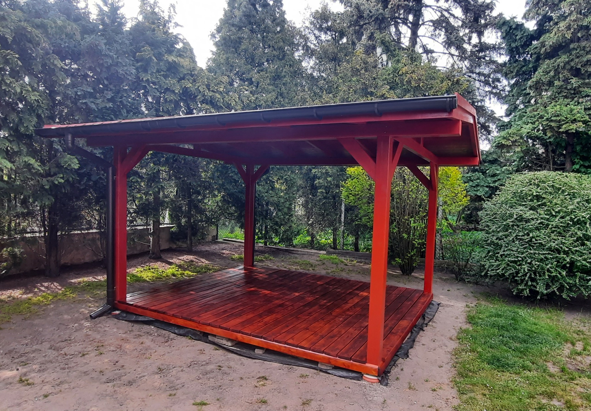 Czerwona, drewniana konstrukcja z dachem i podłogą, otoczona zielenią ogrodu, z widocznym systemem rynnowym.