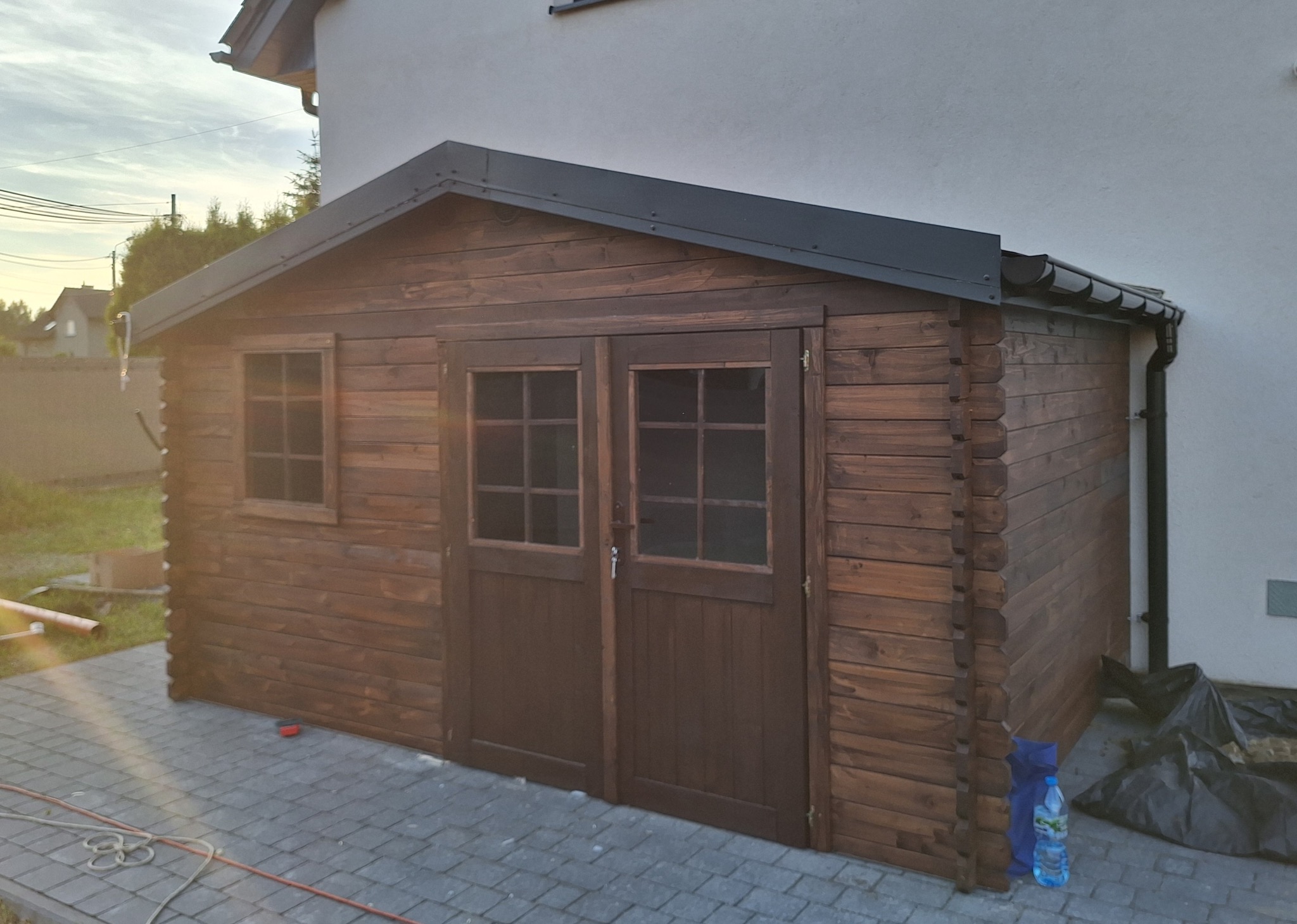 Drewniana konstrukcja z dwuskrzydłowymi drzwiami i małym oknem, przylegająca do budynku z szarą elewacją, otoczona kostką brukową, z widocznymi elementami wyposażenia i butelką wody.
