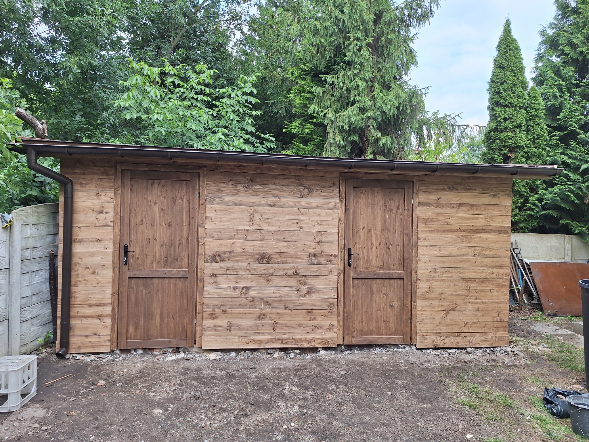 Nowo wybudowana drewniana altana z dwiema parami drzwi, widok z zewnątrz w ogrodzie z widocznym systemem rynnowym.