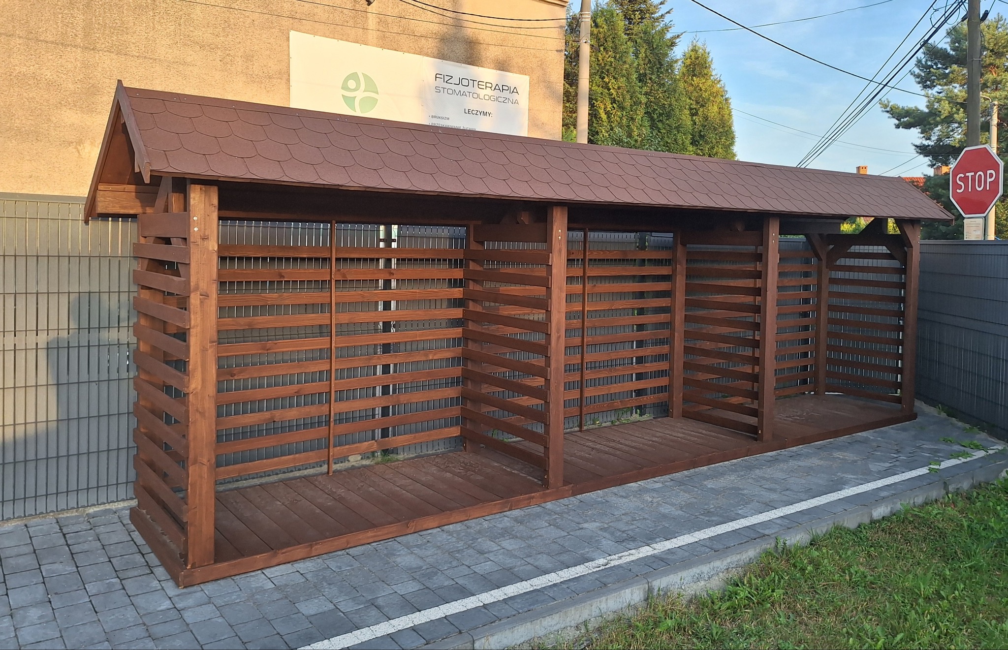 Brązowa drewniana konstrukcja z dachem z gontu bitumicznego, podzielona na trzy segmenty, z poziomymi listwami i metalową siatką, stojąca na kostce brukowej przed budynkiem z szyldem gabinetu...