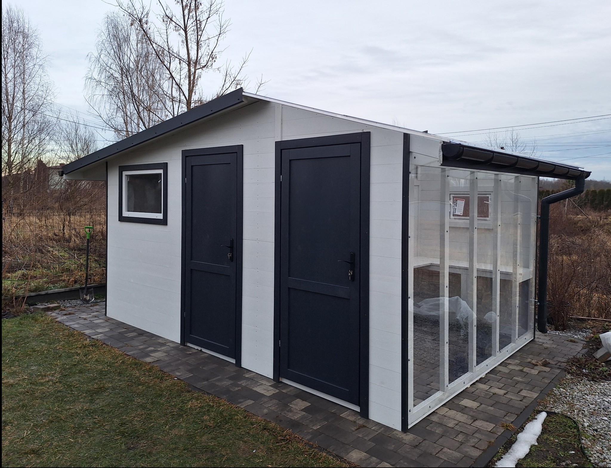 Nowoczesna altana ogrodowa z białymi ścianami, dwiema ciemnymi drzwiami i przezroczystym panelem bocznym, otoczona trawnikiem i kostką brukową.