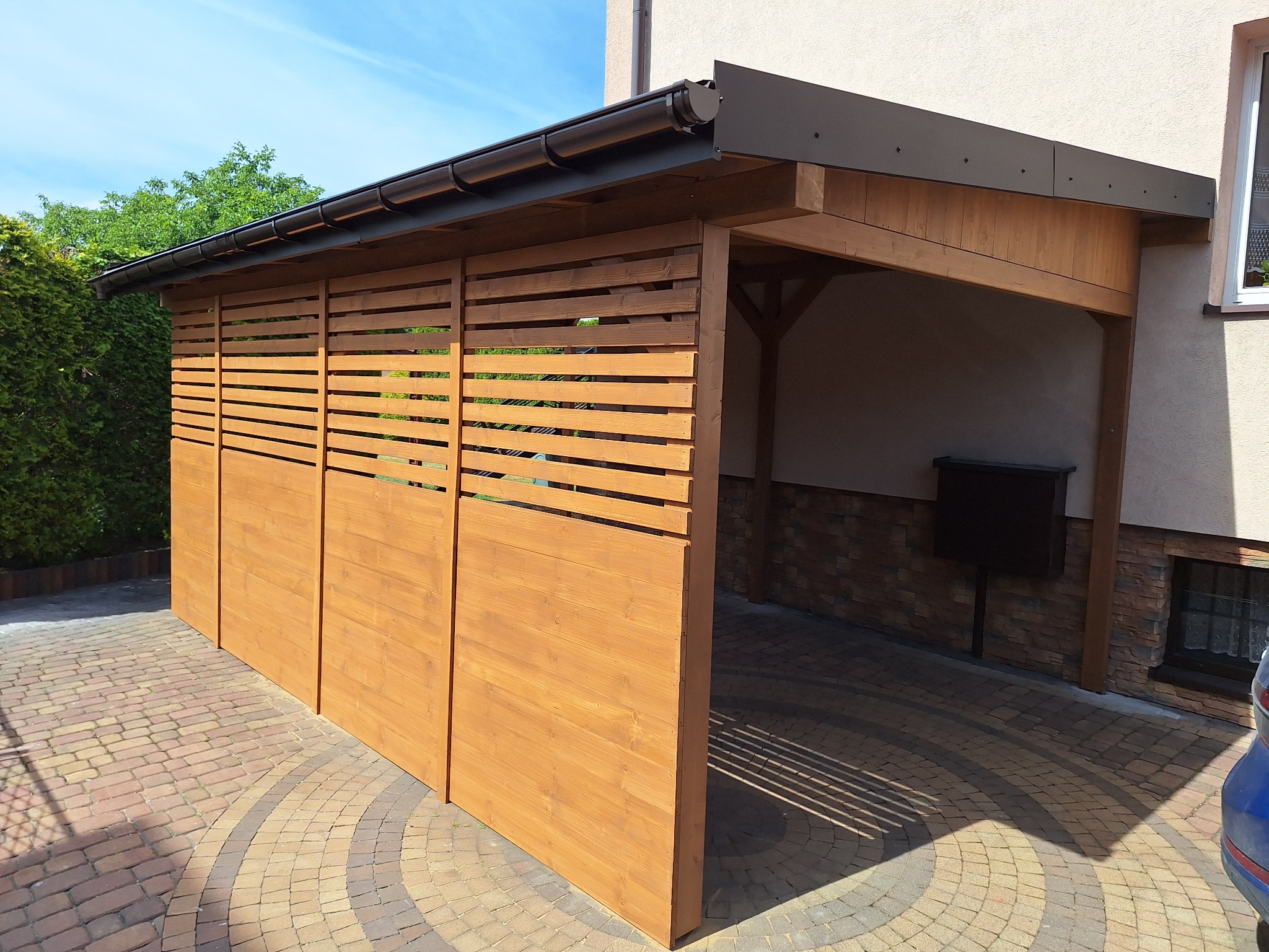 Drewniana konstrukcja z poziomymi szczeblami, prawdopodobnie wiata lub altana przylegająca do budynku, z widocznym fragmentem kostki brukowej i zieleni w tle.