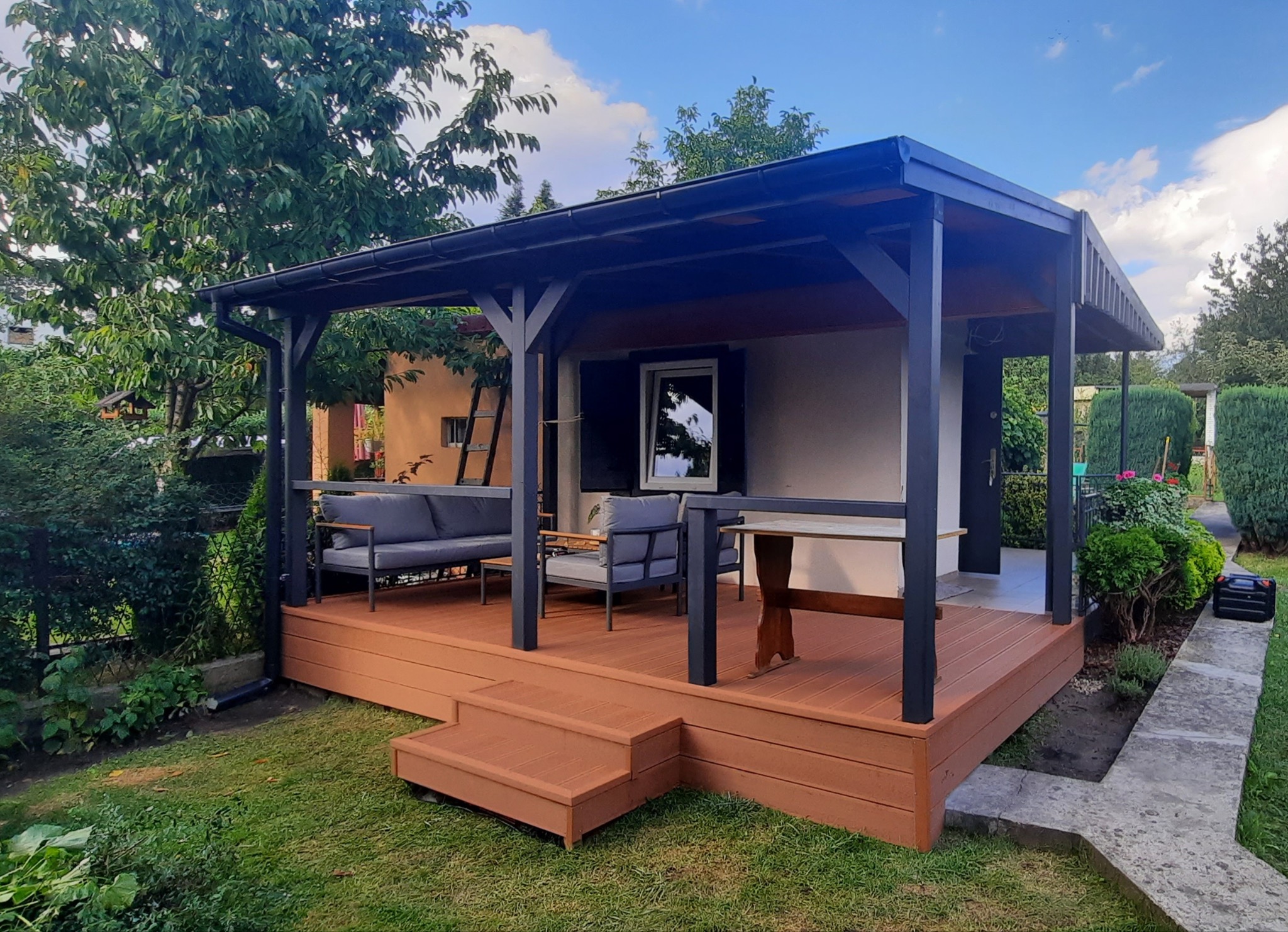 Zadaszony taras z drewnianą podłogą i czarną konstrukcją, wyposażony w szarą sofę i krzesła, z widoczną drabinką opartą o ścianę budynku w tle.