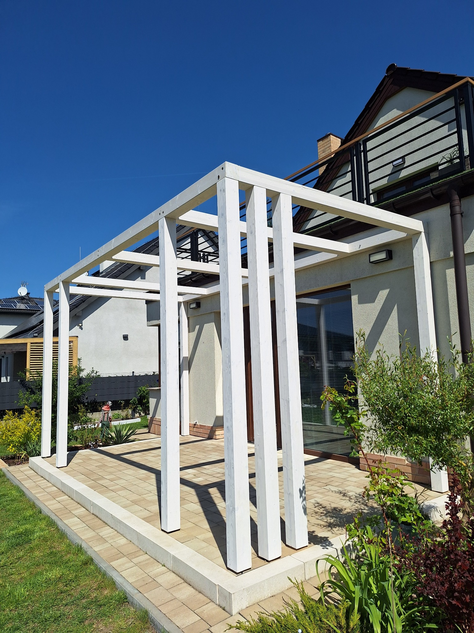 Biała, drewniana pergola na tarasie z kostki, w tle dom z balkonem i bujna roślinność. Słoneczny dzień, błękitne niebo. Widok na ogród i sąsiedni dom.