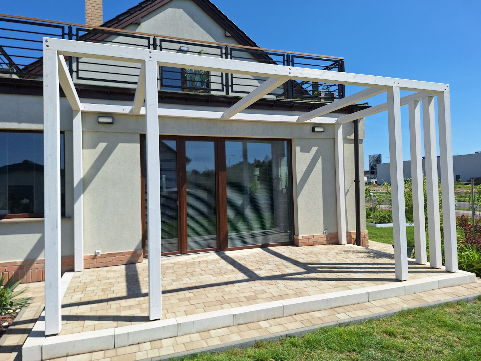 Biała pergola z drewnianą konstrukcją na tarasie z kostki brukowej przy domu z balkonem. Błękitne niebo w tle.