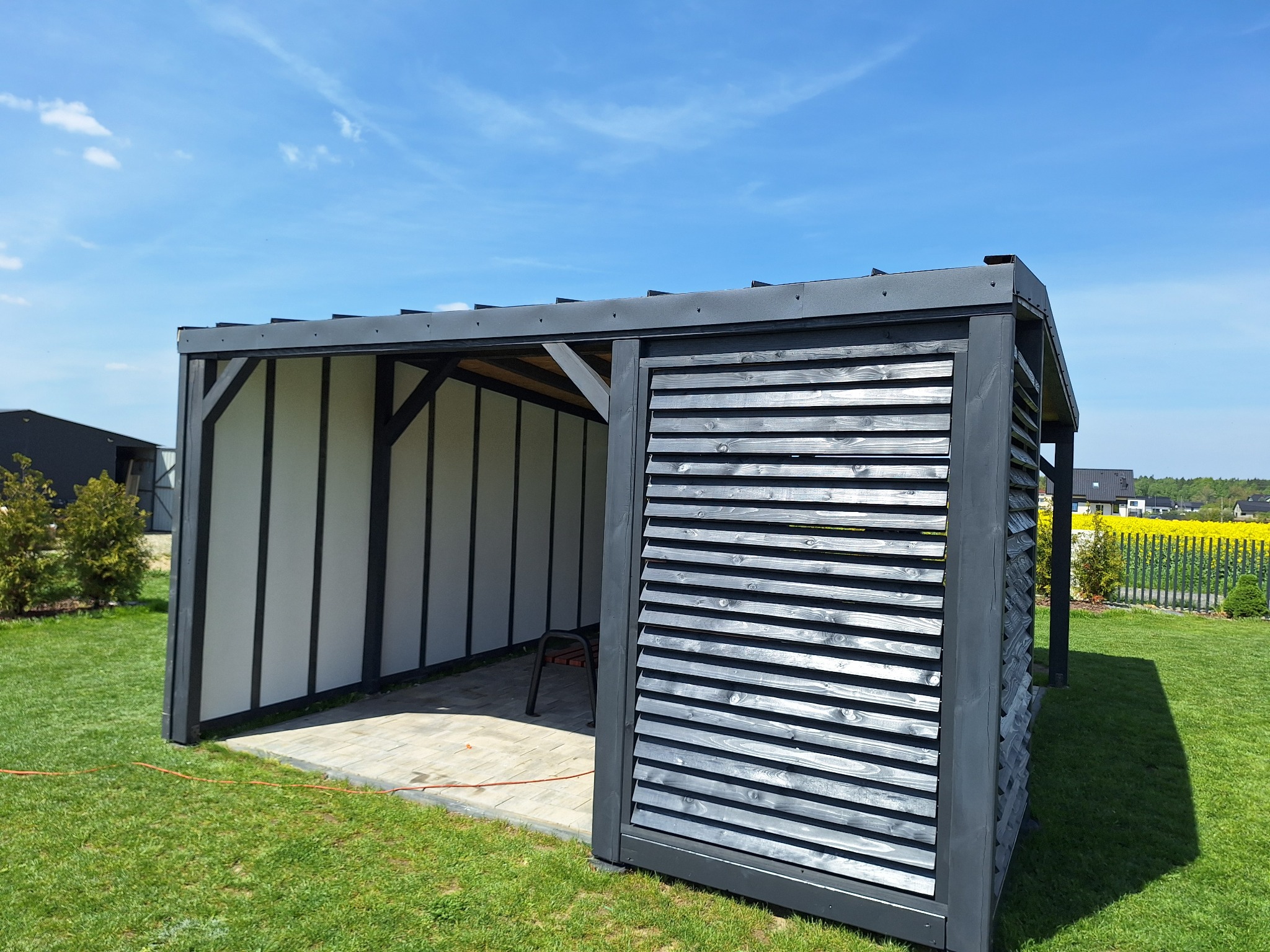 Nowoczesna, grafitowa pergola z drewnianymi lamelami, ustawiona na kostce brukowej w ogrodzie. W tle zielony trawnik i kwitnące pole rzepaku pod błękitnym niebem.