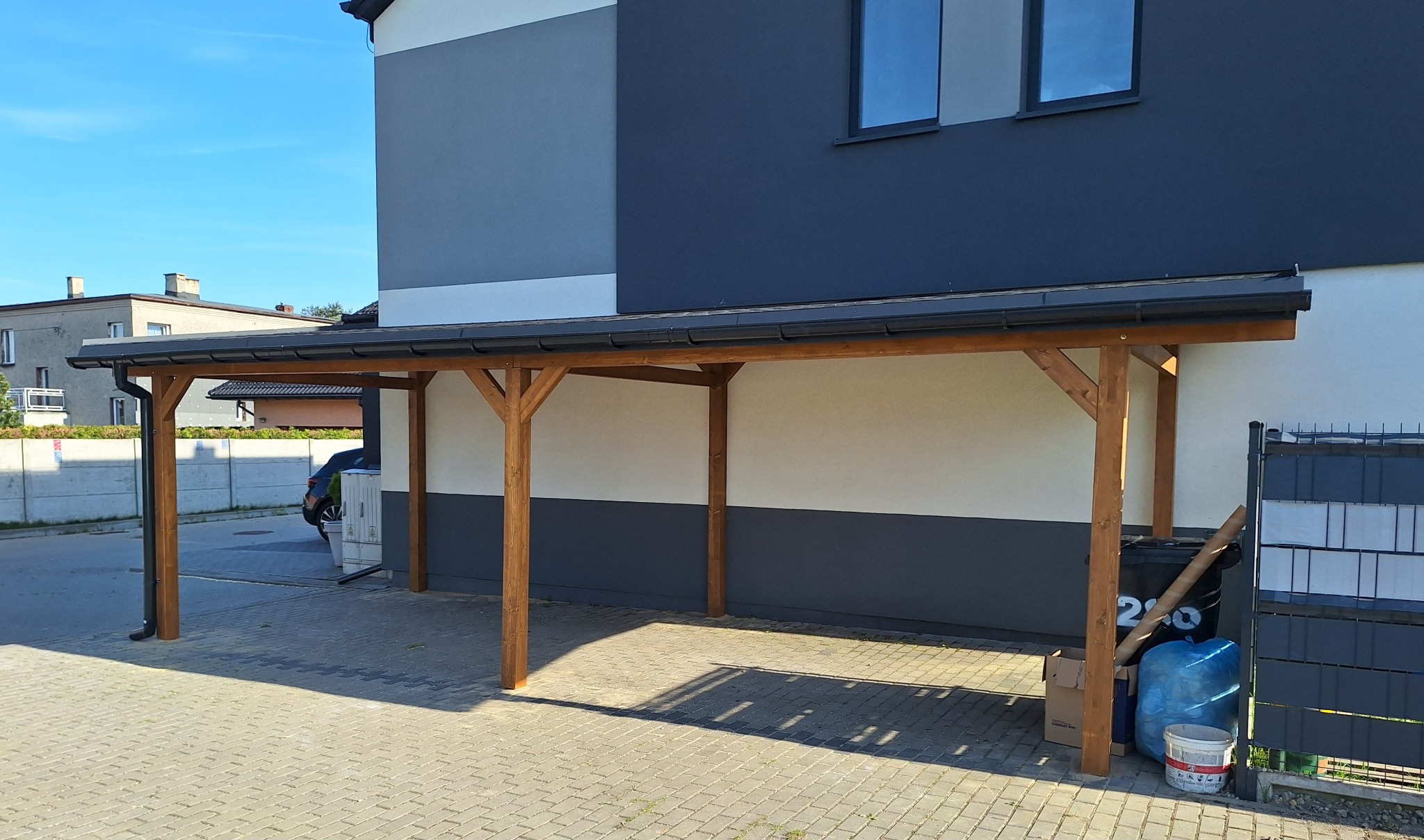 Drewniana pergola przy nowoczesnym domu z szarą elewacją. Konstrukcja z belek, widoczne elementy montażowe. Podłoże z kostki brukowej, w tle zabudowa i zieleń.