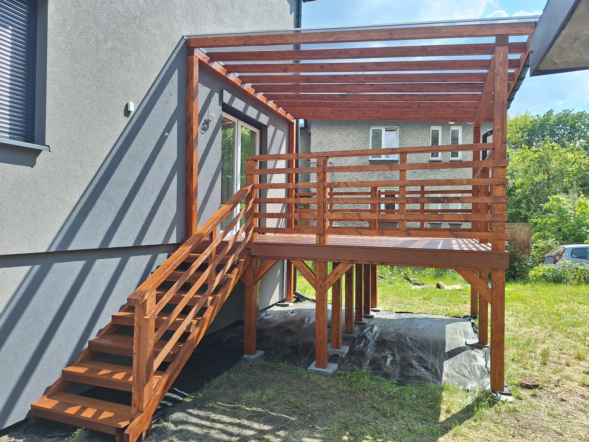 Drewniany taras z zadaszeniem i schodami przy szarym budynku, widok z zewnątrz w słoneczny dzień. Konstrukcja z belek, barierki z desek, zielona trawa.