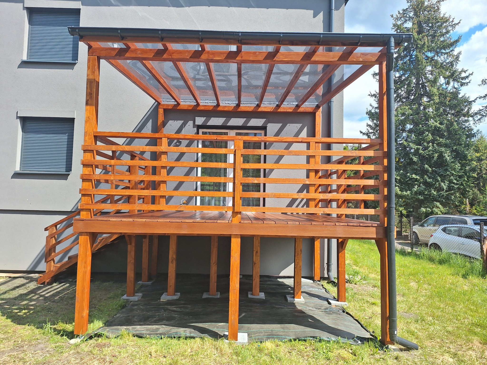 Drewniany taras z zadaszeniem z przezroczystego tworzywa, schody z balustradą, szare ściany budynku i zieleń drzew w tle, w słoneczny dzień.