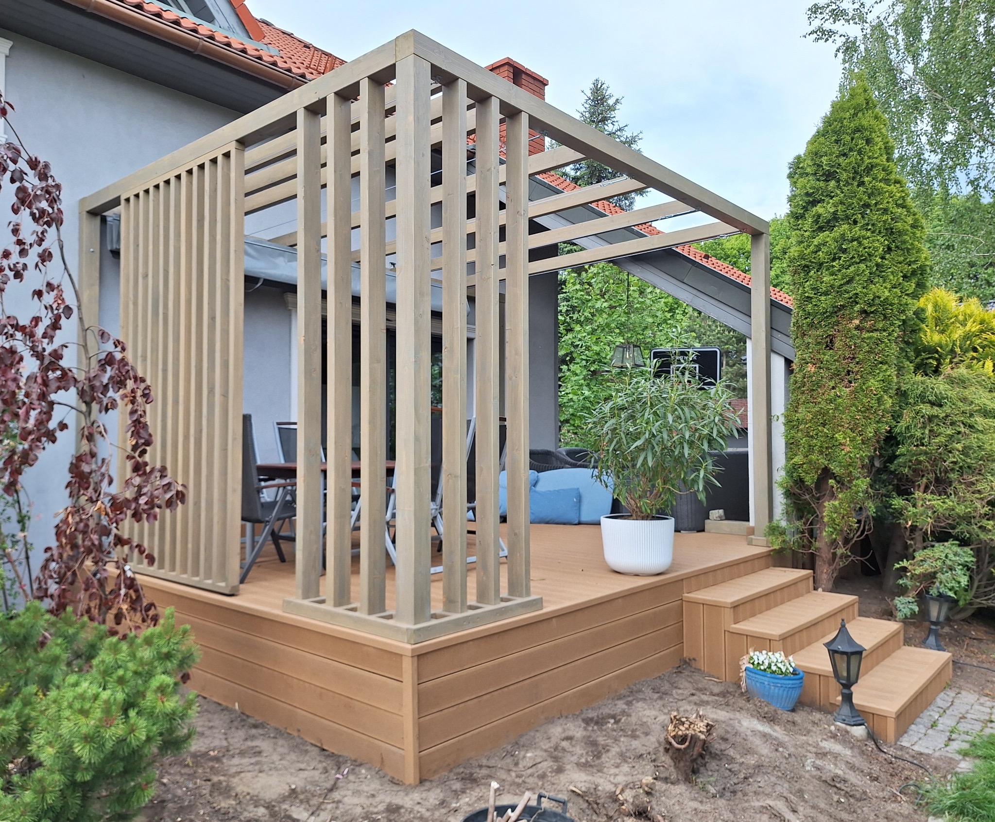 Nowoczesna pergola z drewnianą konstrukcją i tarasem kompozytowym. Widoczne meble ogrodowe i dekoracyjne rośliny. Całość wkomponowana w zielony ogród.