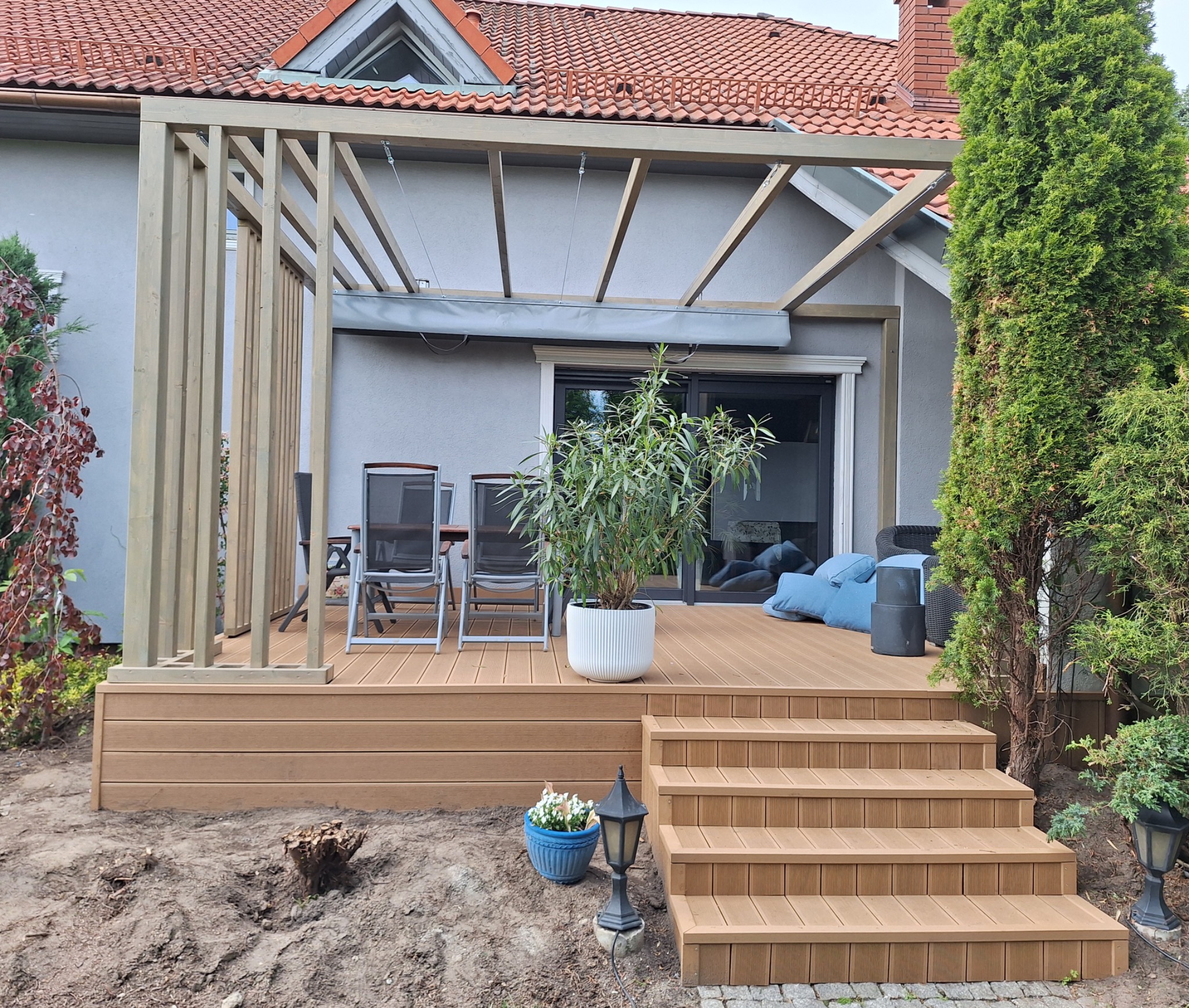 Kompozytowy taras z pergolą i schodami, jasne drewno, widok na dom i ogród. Elementy dekoracyjne: rośliny w donicach, lampa ogrodowa. Stonowana kolorystyka.