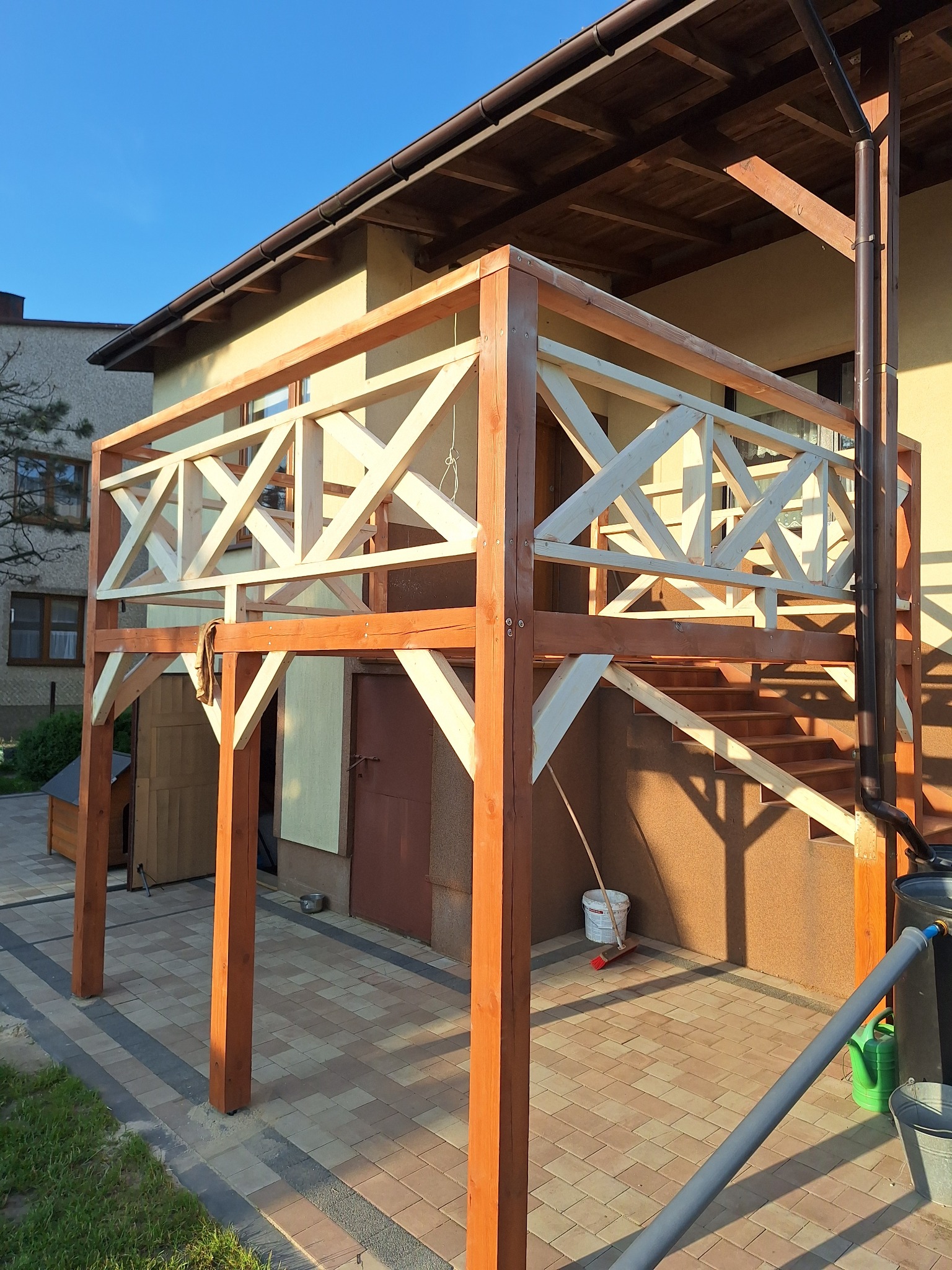 Wznoszona drewniana konstrukcja tarasu przy budynku z widocznymi schodami i balustradą z krzyżowych elementów. Częściowo pomalowane elementy i narzędzia w tle.