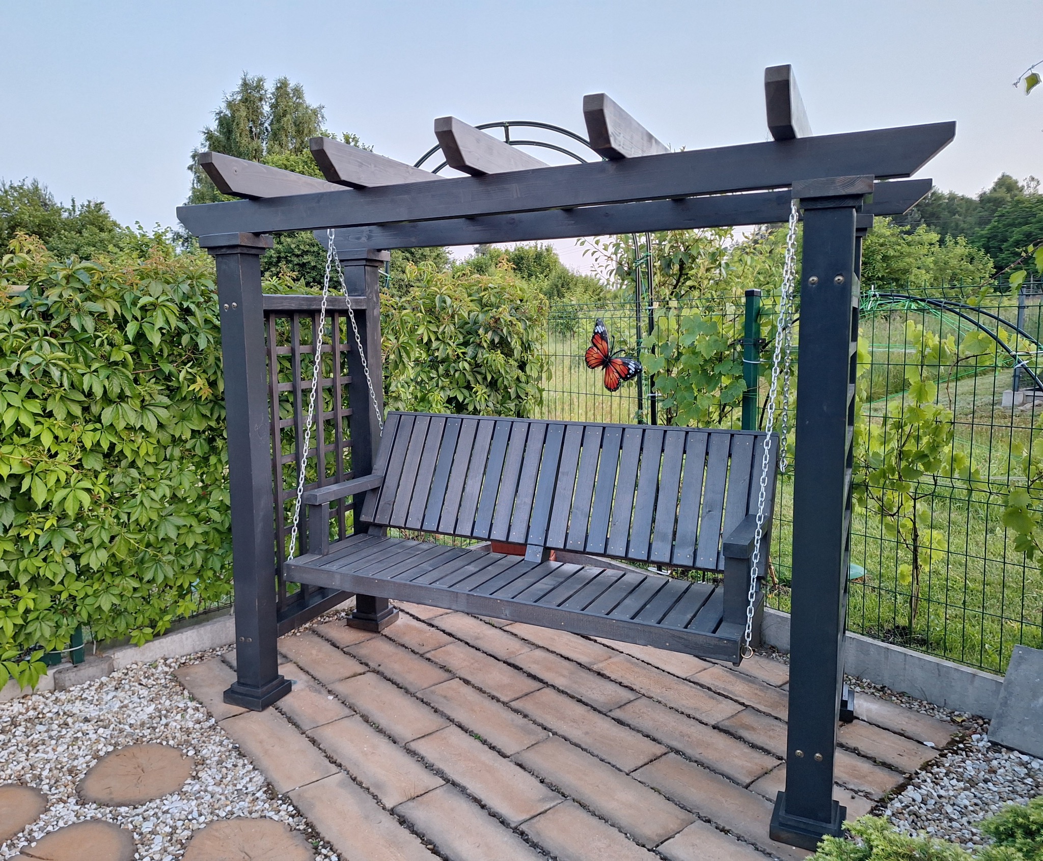 Ciemna, drewniana pergola z huśtawką na tarasie z kostki, otoczona zielenią i dekoracją motyla. Widok na ogród w tle.