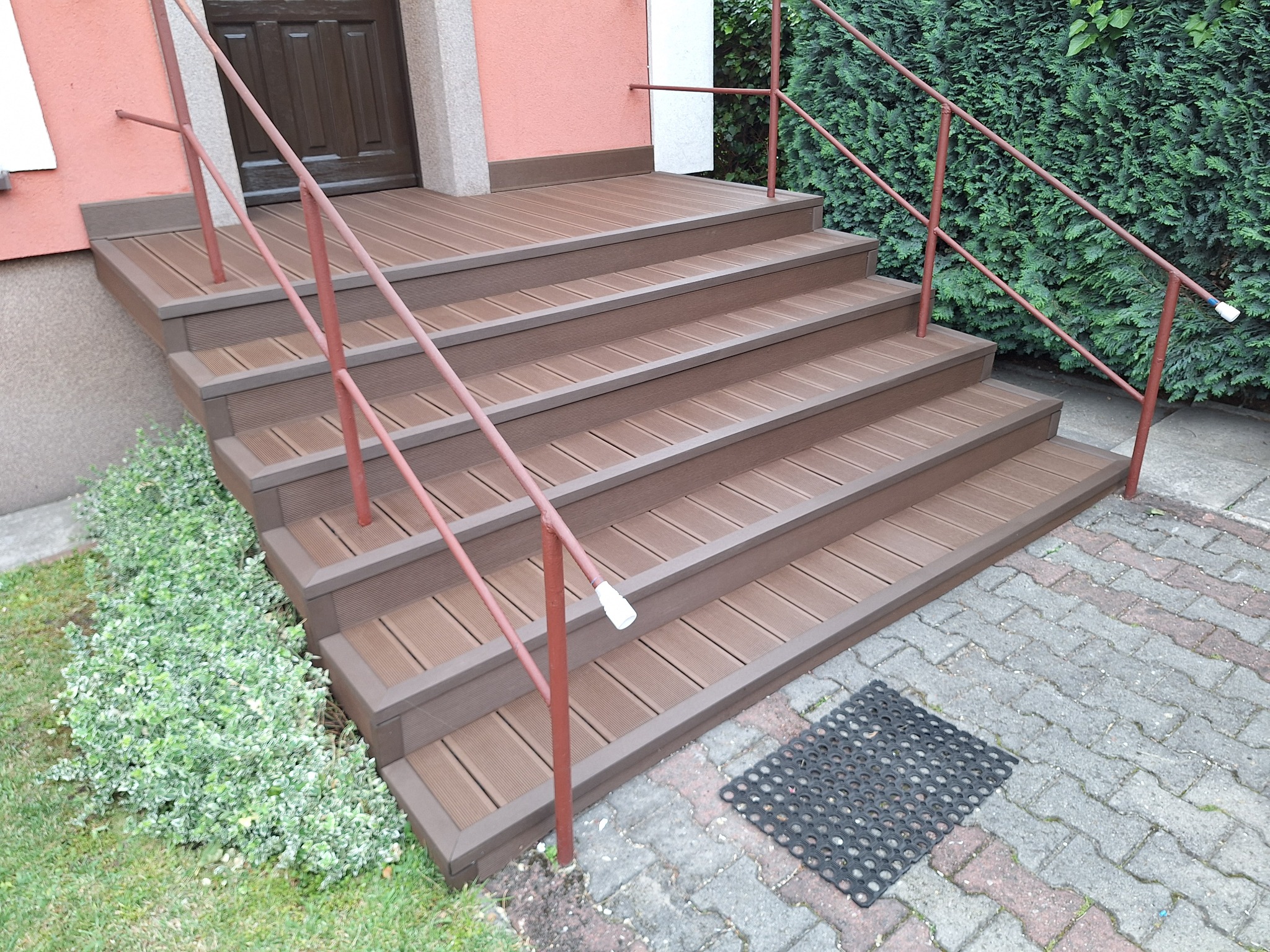 Schody zewnętrzne z brązowych desek kompozytowych prowadzące do drzwi, z poręczą w kolorze ceglastym. Kostka brukowa, zielona roślinność w tle.