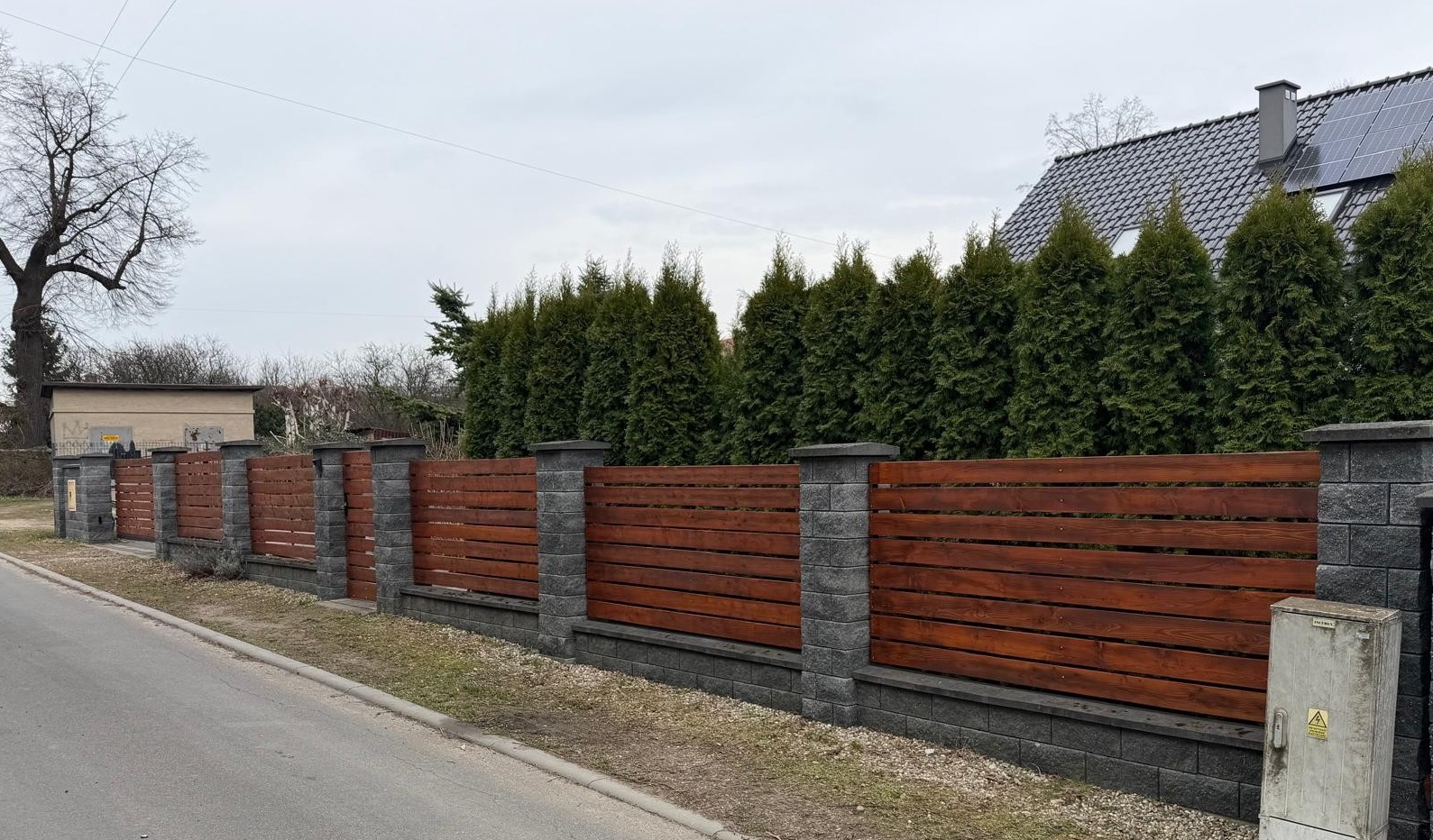 Nowoczesne ogrodzenie z drewnianymi panelami i kamiennymi słupkami, otoczone zielenią, z domem w tle. Widok z poziomu ulicy, pochmurny dzień.