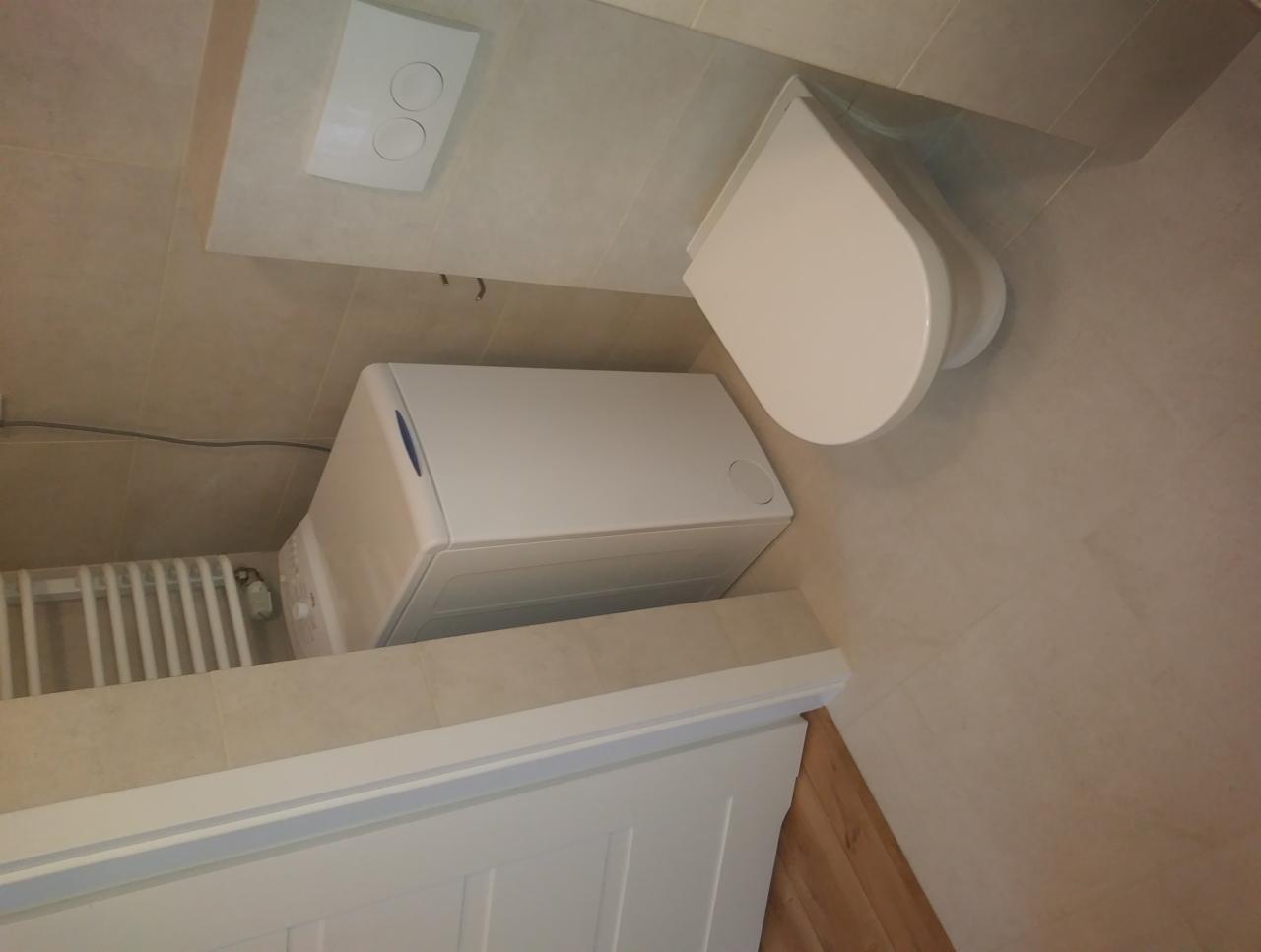 Ukończona łazienka z beżowymi płytkami, podwieszana toaleta, pralka ładowana od góry oraz grzejnik drabinkowy.