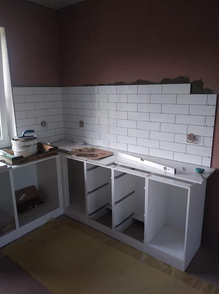 Wnętrze kuchni w trakcie montażu: białe, minimalistyczne szafki bez frontów, częściowo ułożone białe, prostokątne płytki na ścianie, poziomica na blacie, wiadro z zaprawą przy oknie.