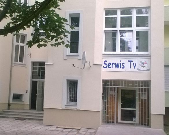 Fasada budynku z napisem 'Serwis Tv' nad wejściem, antena satelitarna obok, okratowane okno i drzwi.