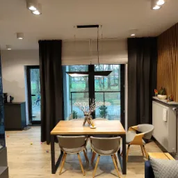 Jadalnia z drewnianym stołem na metalowych nogach, szarymi krzesłami i dekoracyjnym bukietem w wazonie, widok na zewnątrz przez okno z roletami rzymskimi.