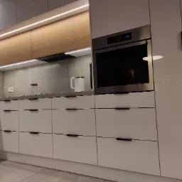 Nowoczesna kuchnia z białymi, lakierowanymi frontami szafek, drewnianą górną zabudową z oświetleniem LED, szarym blatem i wbudowanym piekarnikiem.