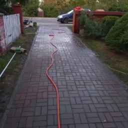 Świeżo ułożona, mokra kostka brukowa w odcieniach brązu, z widocznym wężem ogrodowym, prowadząca do bramy wjazdowej i zaparkowanego samochodu.