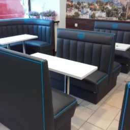 Ustawienie czarnych, tapicerowanych boksów z jasnoniebieską lamówką i białymi stolikami w lokalu gastronomicznym, widok z perspektywy siedzącego.