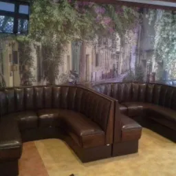 Brązowe, skórzane meble tapicerowane w kształcie półkola, z wysokimi oparciami, ustawione w lokalu z fototapetą przedstawiającą uliczkę miejską.