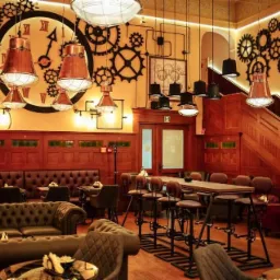 Wnętrze lokalu w stylu steampunk: drewniane panele, sofa Chesterfield, wysokie krzesła barowe z motywem przekładni, dekoracje ścienne z zębatek i zegara, miedziane lampy wiszące.