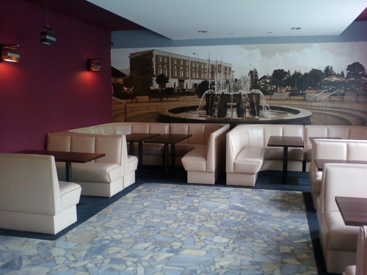 Wnętrze lokalu gastronomicznego z tapicerowanymi narożnikami w jasnym odcieniu, ciemnymi stolikami i mozaikową podłogą, na ścianie fototapeta z fontanną.