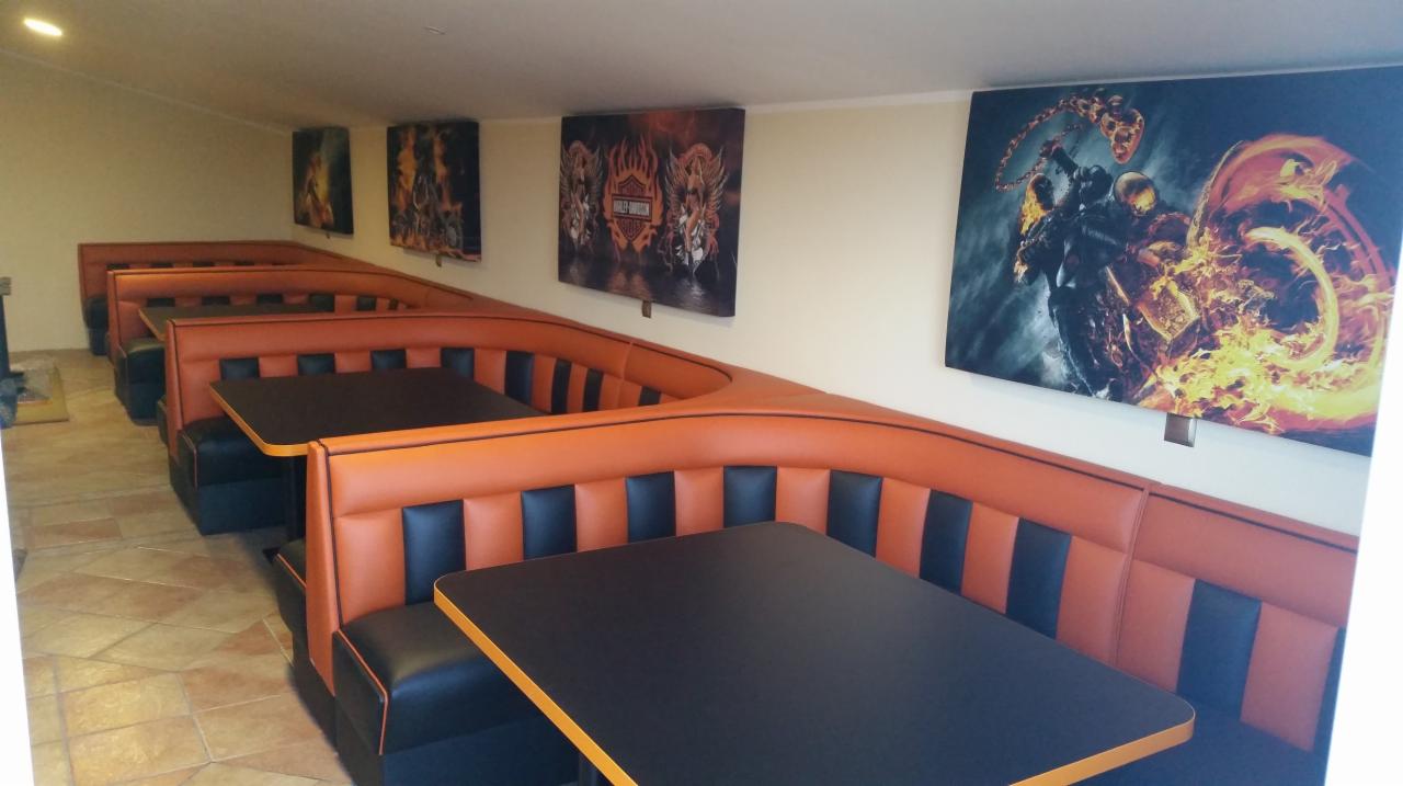 Rząd tapicerowanych, pomarańczowo-czarnych boksów restauracyjnych z czarnymi blatami, pod ścianą z obrazami motocyklowymi.