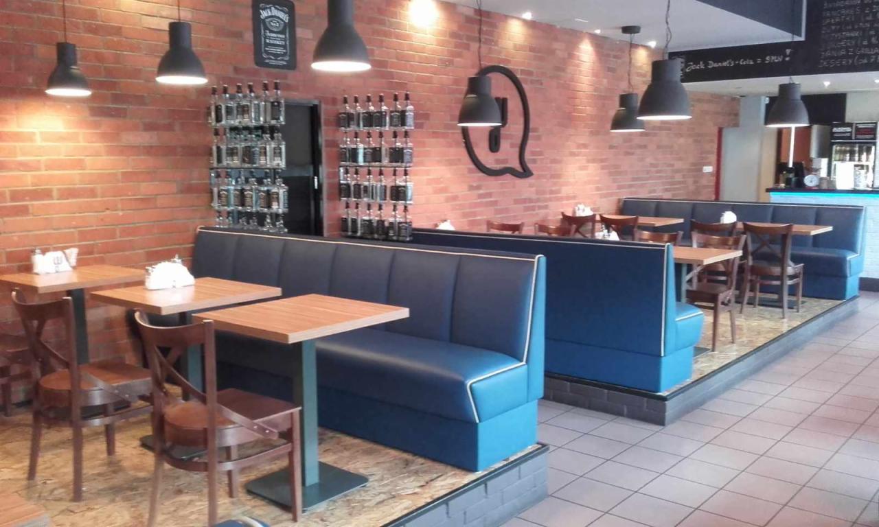Wnętrze restauracji z ceglaną ścianą, ciemnymi lampami wiszącymi nad drewnianymi stolikami i niebieskimi tapicerowanymi lożami z białymi lamówkami. Na ścianie dekoracja w kształcie dymka i półki...