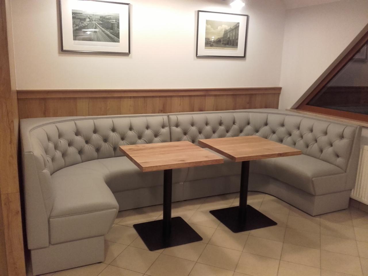 Szara, narożna, pikowana kanapa z dwoma drewnianymi stolikami na czarnych, kwadratowych podstawach, ustawiona w lokalu z drewnianą boazerią i obrazami na ścianach.