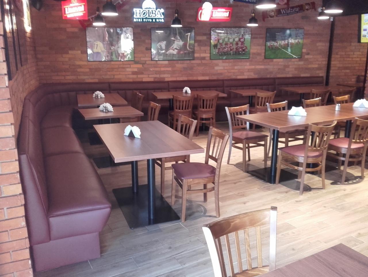 Wnętrze lokalu gastronomicznego z długą, tapicerowaną ławą w kolorze burgundzkim, drewnianymi stołami z czarnymi nogami i krzesłami, ściany z cegły, dekoracje sportowe i oświetlenie punktowe.