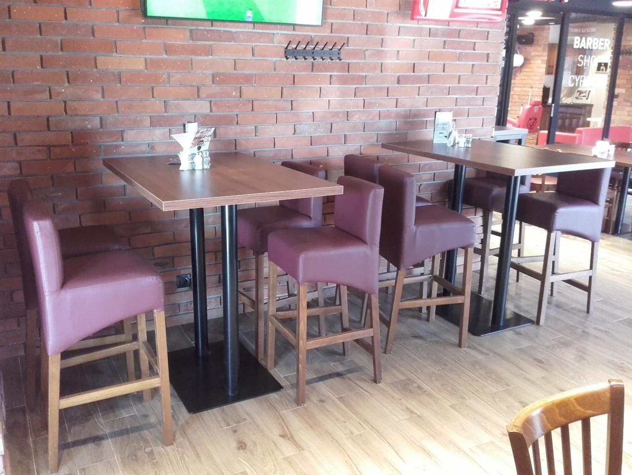 Wysokie krzesła barowe z bordową tapicerką i drewnianymi nogami ustawione przy kwadratowych stolikach z ciemnymi blatami na tle ściany z czerwonej cegły w lokalu gastronomicznym.