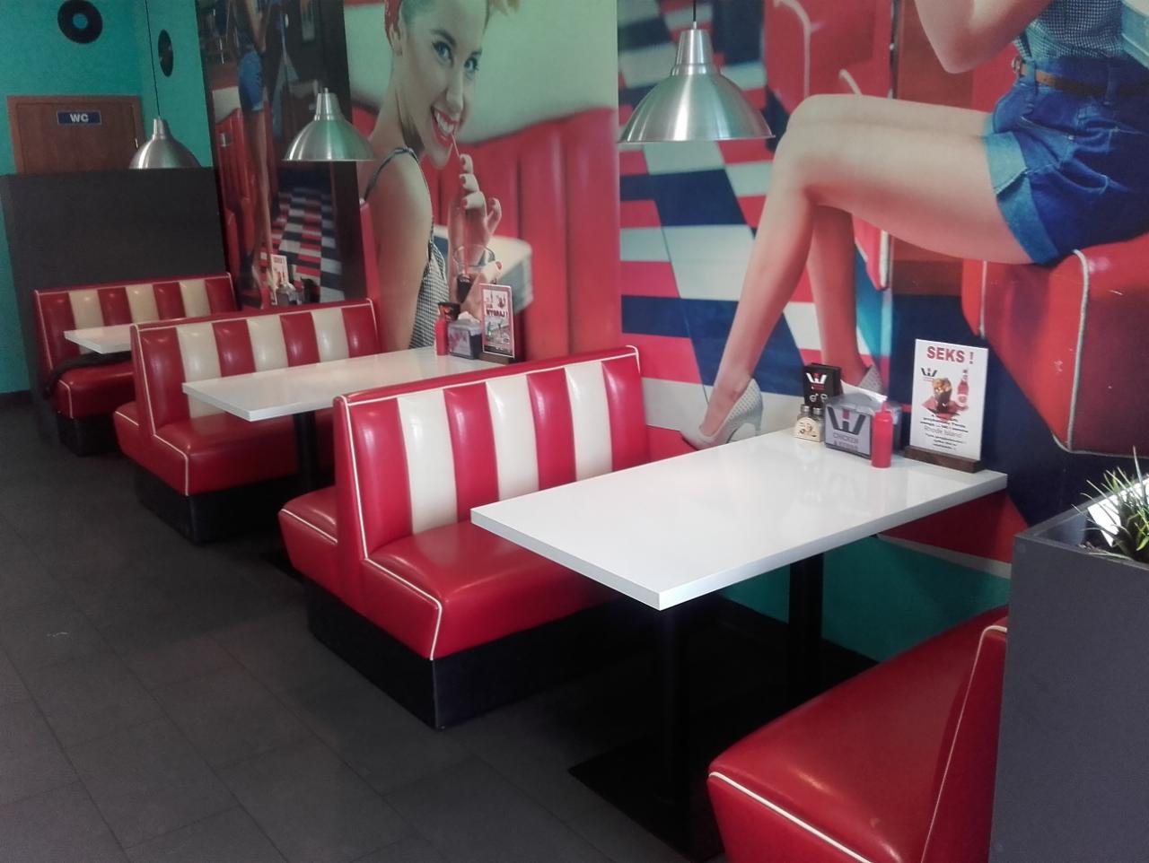 Wnętrze restauracji w stylu retro z czerwono-białymi boksami i białymi stolikami, ozdobione dużym muralem z wizerunkiem kobiety i nóg w szpilkach. Na stoliku menu i przyprawy.