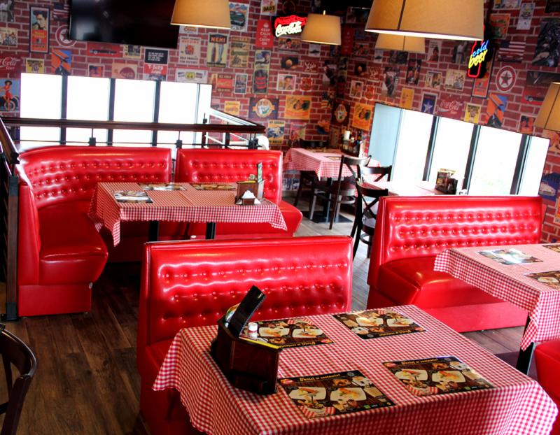 Wnętrze restauracji w stylu amerykańskim: czerwone, tapicerowane boksy z pikowanymi oparciami, stoliki nakryte obrusami w czerwoną kratę, ściany wyłożone cegłą z plakatami retro, lampy wiszące...