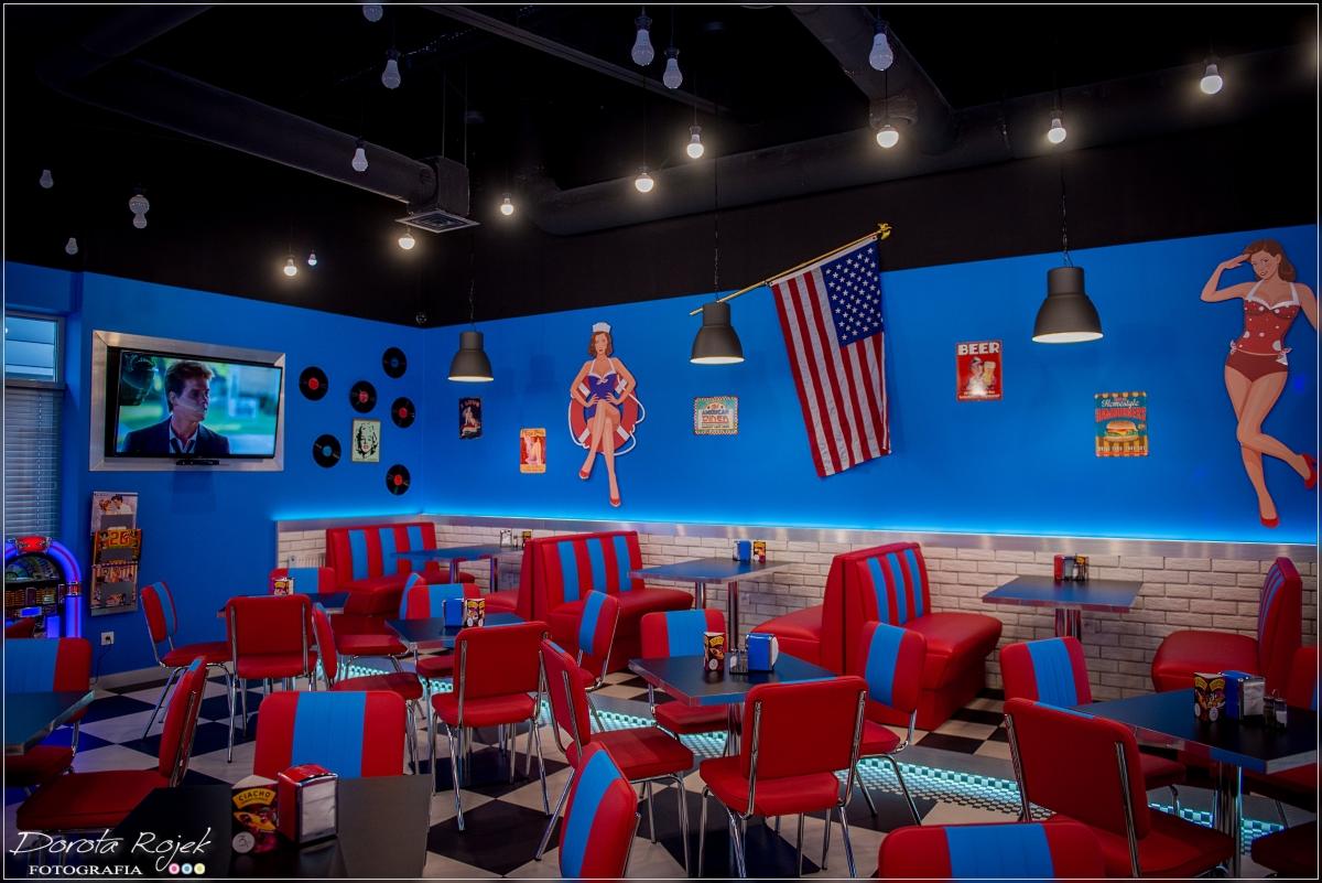 Wnętrze restauracji w stylu amerykańskim z lat 50-tych: niebieskie ściany ozdobione plakatami, flagą USA i sylwetkami pin-up girls, czerwono-niebieskie loże i krzesła, czarno-biała podłoga...