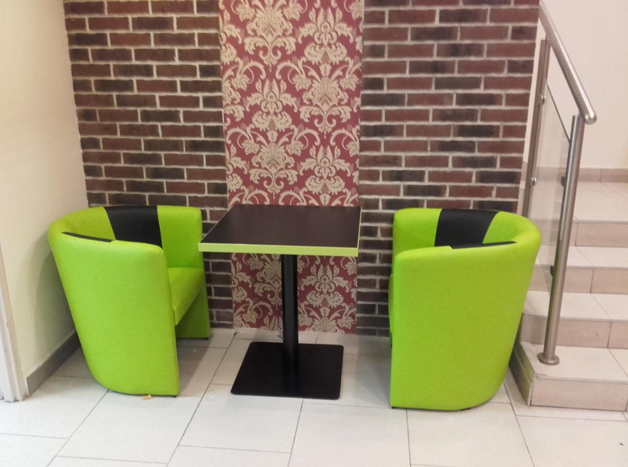 Dwa jasnozielone, tapicerowane fotele z czarnymi oparciami i kwadratowy stolik z czarnym blatem i podstawą, ustawione na białej podłodze w pomieszczeniu z ceglaną ścianą i ozdobną tapetą.