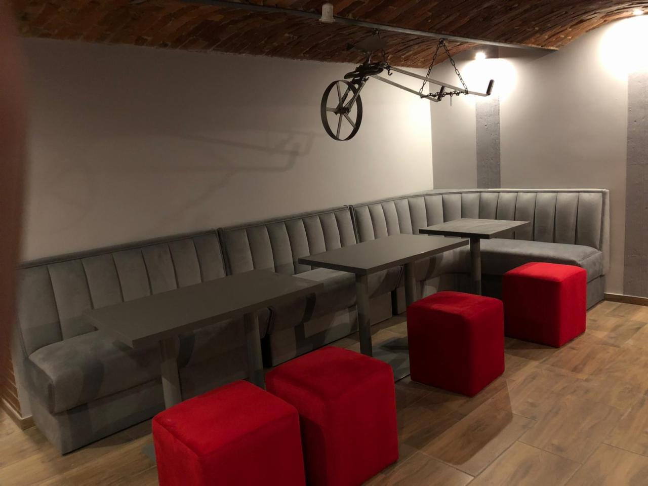Szare, tapicerowane siedzisko narożne z pionowymi przeszyciami, uzupełnione ciemnymi stolikami i czerwonymi pufami na drewnianej podłodze, pod ceglanym sklepieniem z industrialną lampą.