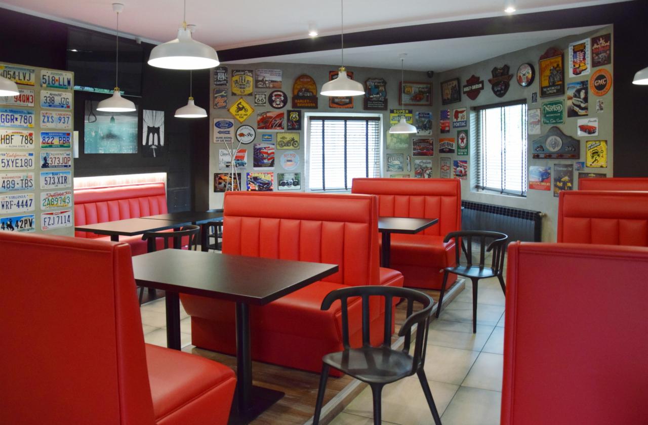 Wnętrze lokalu gastronomicznego w stylu retro z czerwonymi lożami i stolikami oraz ścianami ozdobionymi tablicami rejestracyjnymi i plakatami motoryzacyjnymi.