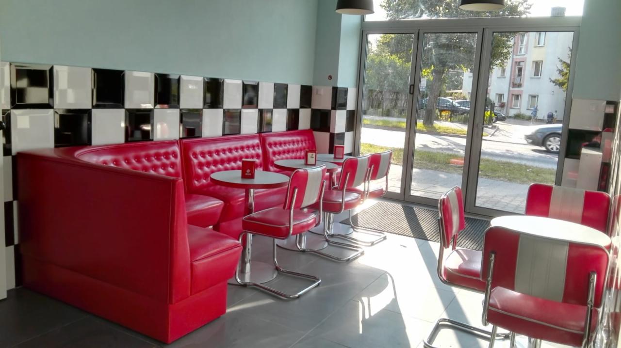 Wnętrze lokalu w stylu retro: czerwone, pikowane loże i krzesła ze srebrnymi nogami, okrągłe stoliki, ściana w czarno-białą szachownicę, przeszklone drzwi wejściowe z widokiem na ulicę.