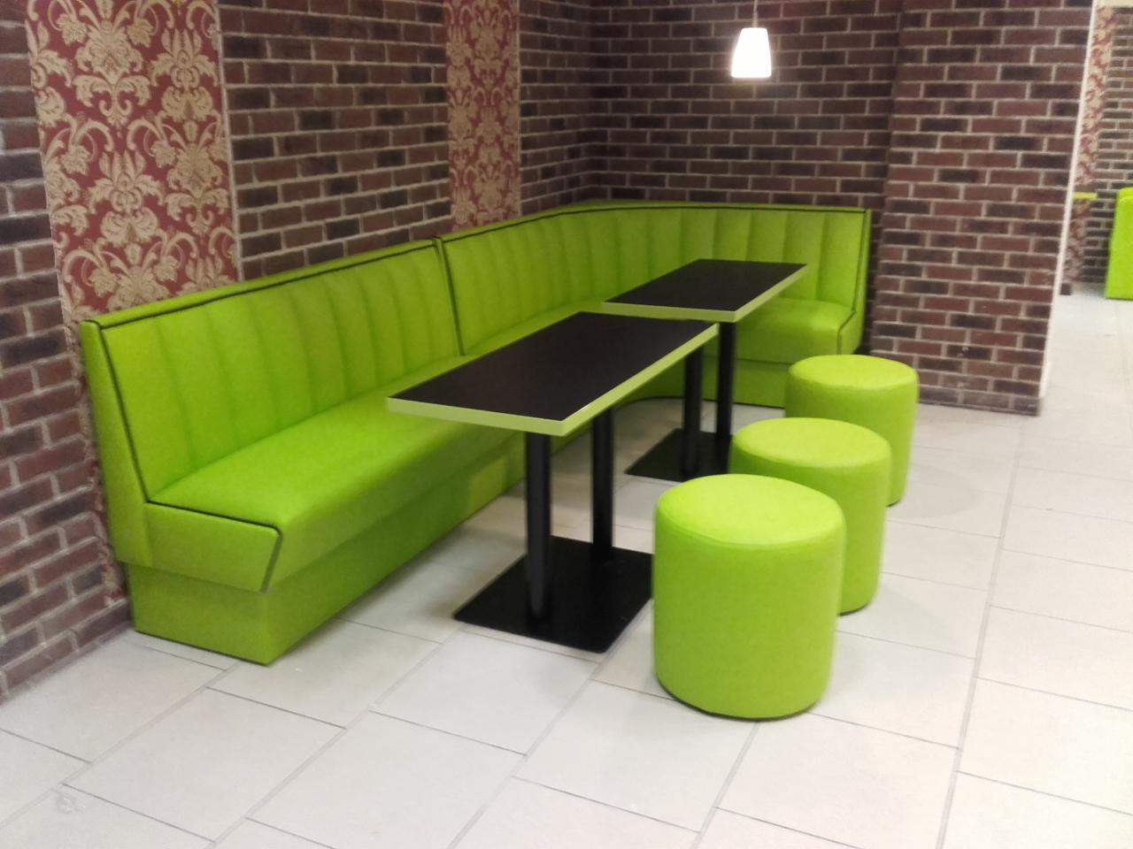 Jasnozielony narożnik tapicerowany z czarną lamówką, stoliki z czarnymi blatami i okrągłe pufy w lokalu z ceglanymi ścianami i wzorzystą tapetą.